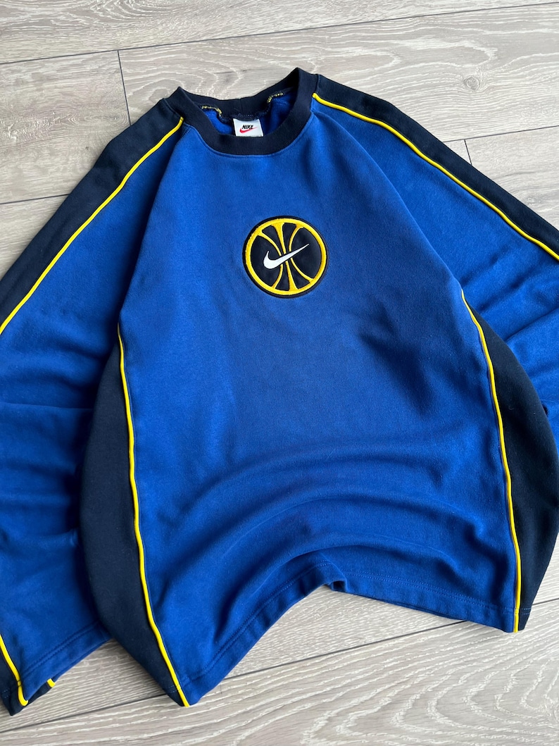 May include: A blue sweatshirt with a round Nike logo featuring a basketball design. The sweatshirt has black and yellow accents on the sleeves and sides. The Nike logo is centered on the chest.
