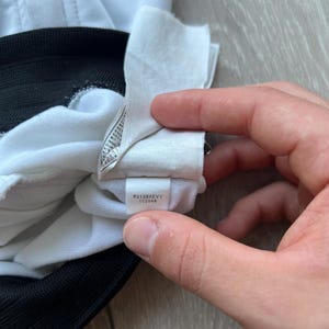 May include: Close-up of a white garment with a black trim. A tag with the text "F31204EVY 112049" is visible. The garment appears to be made of a soft, possibly athletic material.