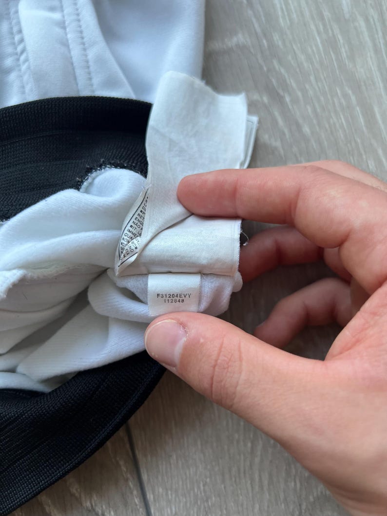 May include: Close-up of a white garment with a black trim. A tag with the text "F31204EVY 112049" is visible. The garment appears to be made of a soft, possibly athletic material.