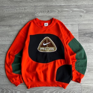 May include: A bright orange crewneck sweatshirt featuring large black color-blocked panels on the front and dark green panels on the sleeves. A triangular patch on the chest displays a white Nike swoosh logo above the letters ACG in white.