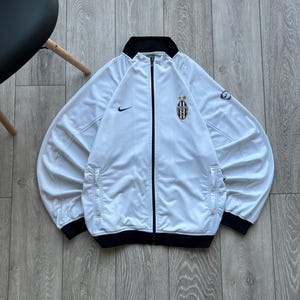 May include: White zip-up track jacket with black trim and a black collar. Features the Nike swoosh logo and a team crest on the chest. The jacket has two front pockets and a full-length zipper.