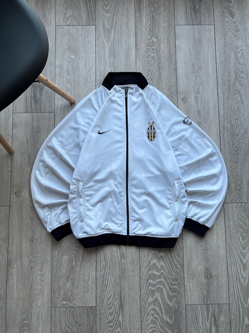 May include: White track jacket with black trim and a black collar. It features the Nike swoosh logo and a team crest on the chest. The jacket has two front pockets and a full-length zip.
