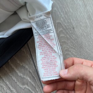 May include: A white tag with black text, attached to a black and white garment. The tag includes text in multiple languages, including English, French, and Spanish, with warnings about fire.