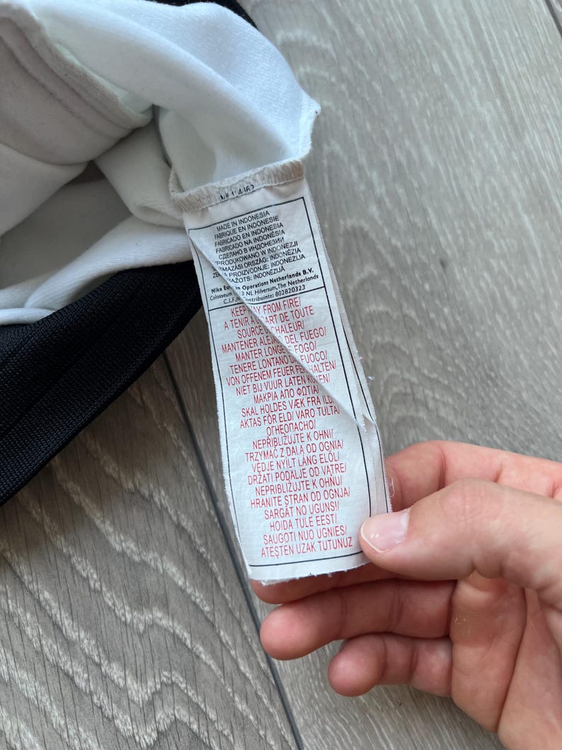 May include: A white tag with black text, attached to a black and white garment. The tag includes text in multiple languages, including English, French, and Spanish, with warnings about fire.