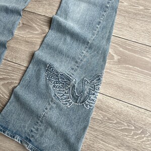 May include: Light blue denim jeans featuring a flared leg silhouette. The lower portion of the leg displays a prominent embroidered design of a horseshoe shape flanked by two detailed wings, executed in a textured, frayed-edge blue thread.