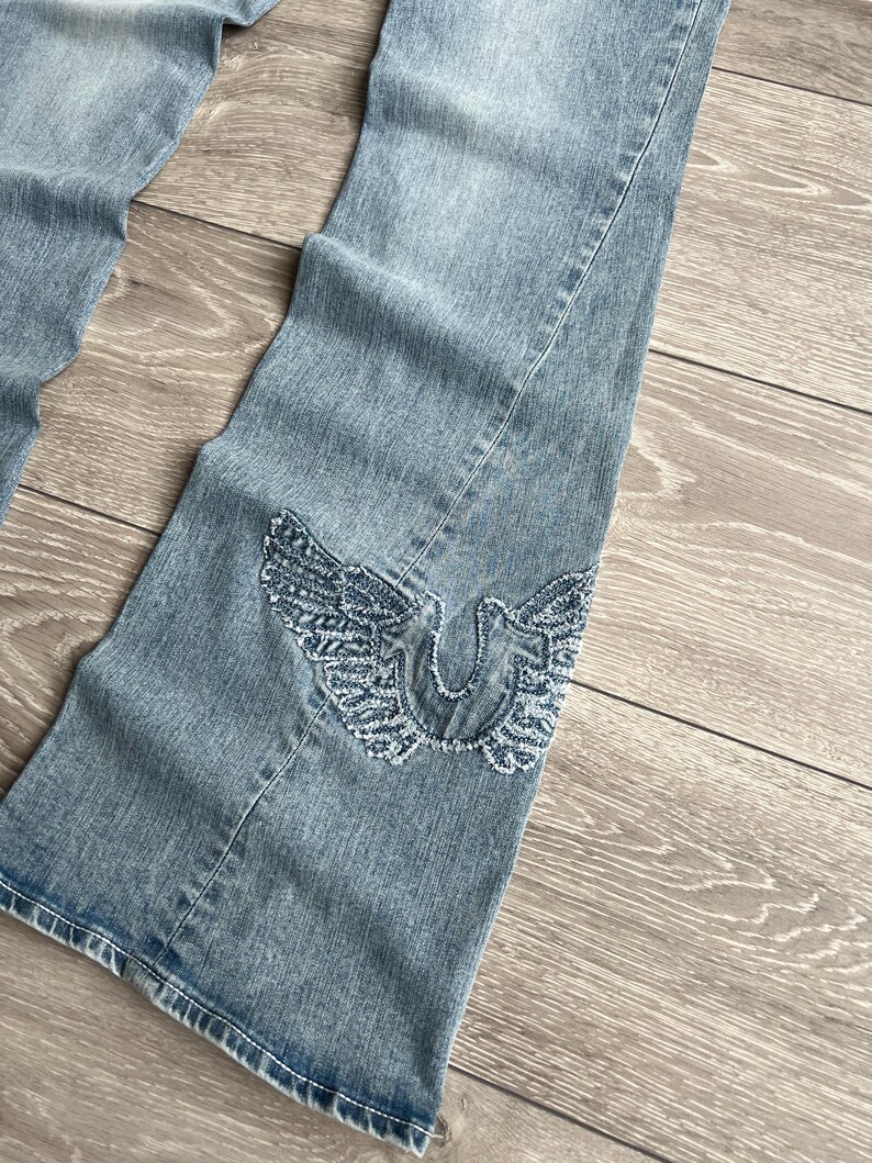 May include: Light blue denim jeans featuring a flared leg silhouette. The lower portion of the leg displays a prominent embroidered design of a horseshoe shape flanked by two detailed wings, executed in a textured, frayed-edge blue thread.