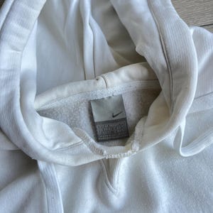 Puede incluir: Primer plano de una sudadera con capucha Nike blanca con cord&oacute;n. La sudadera tiene una franja azul en la parte inferior y un peque&ntilde;o logotipo de Nike en una etiqueta. La tela parece suave y ligeramente desgastada.