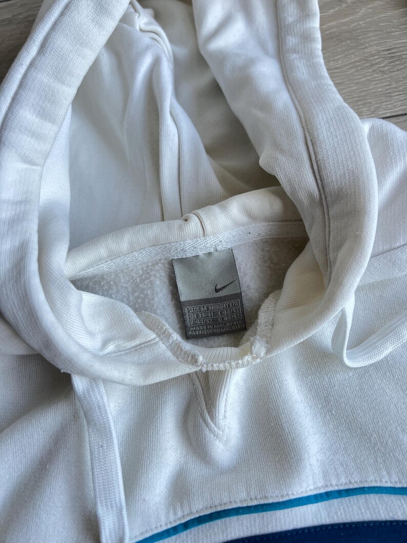 Puede incluir: Primer plano de una sudadera con capucha Nike blanca con cord&oacute;n. La sudadera tiene una franja azul en la parte inferior y un peque&ntilde;o logotipo de Nike en una etiqueta. La tela parece suave y ligeramente desgastada.