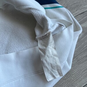 Puede incluir: Sudadera con capucha blanca con un dise&ntilde;o de rayas azules y turquesas en la parte superior del pecho. El interior es de un material suave y afelpado. Una etiqueta est&aacute; adherida al dobladillo inferior.