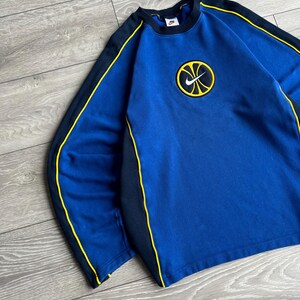 May include: Blue sweatshirt with black and yellow accents. The sweatshirt features a round logo with a yellow basketball design and a black Nike swoosh. The sleeves have black panels with yellow trim. The collar is black.