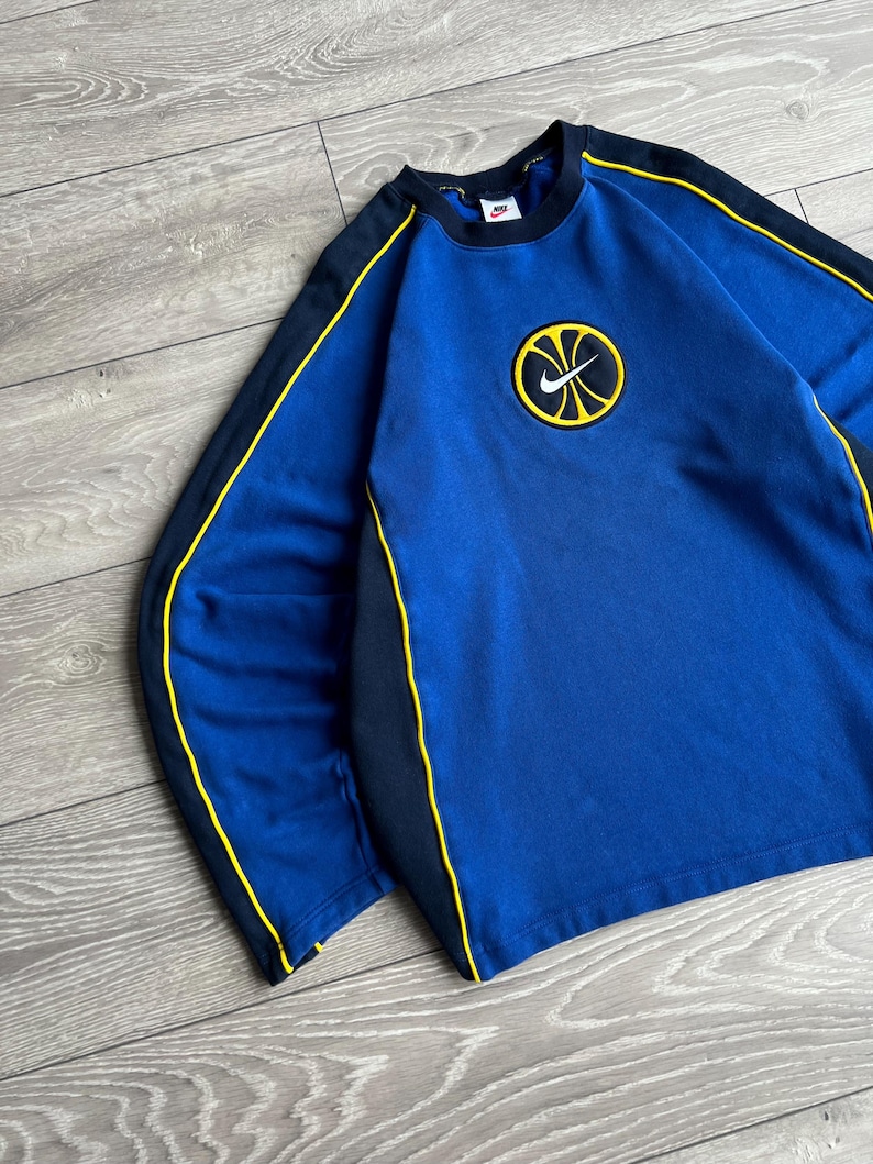 May include: Blue sweatshirt with black and yellow accents. The sweatshirt features a round logo with a yellow basketball design and a black Nike swoosh. The sleeves have black panels with yellow trim. The collar is black.