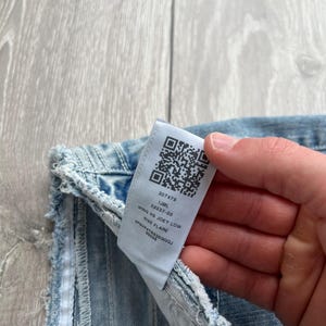 May include: Light blue denim jeans featuring a frayed edge and a white interior care label. The label displays a QR code and the text 207479, IJML, 52037-00, WING HS JOEY LOW, RISE FLARE, and SP6VP4TR838GQQJ 59598 printed in black ink.