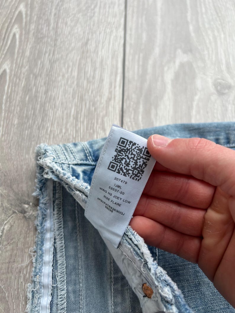 May include: Light blue denim jeans featuring a frayed edge and a white interior care label. The label displays a QR code and the text 207479, IJML, 52037-00, WING HS JOEY LOW, RISE FLARE, and SP6VP4TR838GQQJ 59598 printed in black ink.