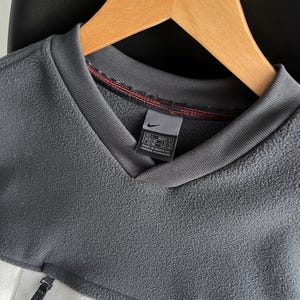 May include: A grey and white fleece pullover with a V-neck collar and a small red Nike logo. The pullover has a zip detail on the side and a tag with size information. The garment is hanging on a wooden hanger.