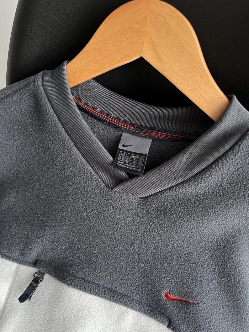 May include: A grey and white fleece pullover with a V-neck collar and a small red Nike logo. The pullover has a zip detail on the side and a tag with size information. The garment is hanging on a wooden hanger.