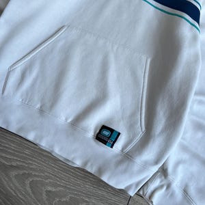 Puede incluir: Sudadera con capucha Nike blanca con un bolsillo delantero y un dise&ntilde;o de rayas azules y turquesas en el pecho. Cuenta con un peque&ntilde;o logotipo de Nike y una etiqueta de EE. UU. La sudadera tiene una capucha con cord&oacute;n y pu&ntilde;os acanalados.