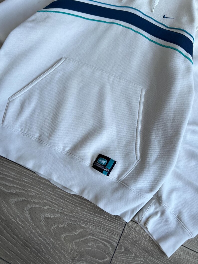 Puede incluir: Sudadera con capucha Nike blanca con un bolsillo delantero y un dise&ntilde;o de rayas azules y turquesas en el pecho. Cuenta con un peque&ntilde;o logotipo de Nike y una etiqueta de EE. UU. La sudadera tiene una capucha con cord&oacute;n y pu&ntilde;os acanalados.