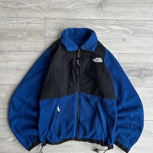 May include: A blue and black fleece jacket with a full zip. The jacket has a blue fleece body with black panels on the upper chest and shoulders. The North Face logo is on the chest.