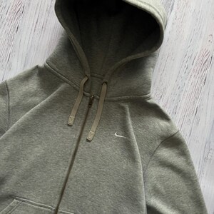 Puede incluir: Sudadera gris con cremallera, capucha con cord&oacute;n y peque&ntilde;o logo blanco de Nike. La sudadera tiene cremallera completa y dos bolsillos delanteros. La tela parece suave y c&oacute;moda.