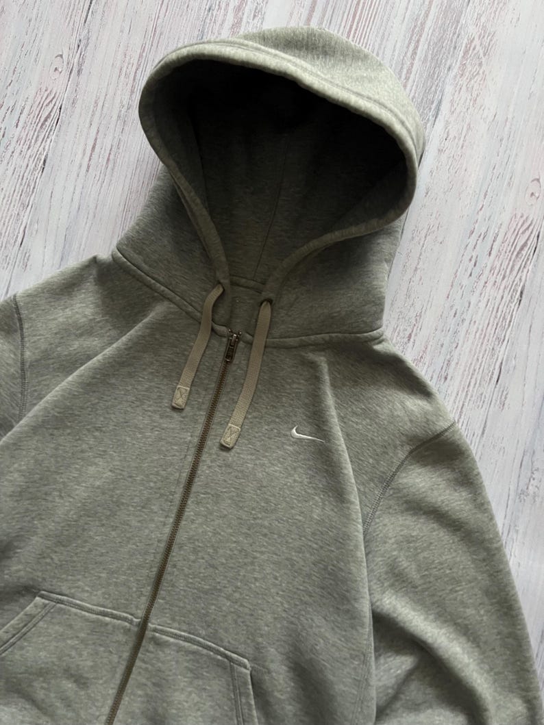 Puede incluir: Sudadera gris con cremallera, capucha con cord&oacute;n y peque&ntilde;o logo blanco de Nike. La sudadera tiene cremallera completa y dos bolsillos delanteros. La tela parece suave y c&oacute;moda.