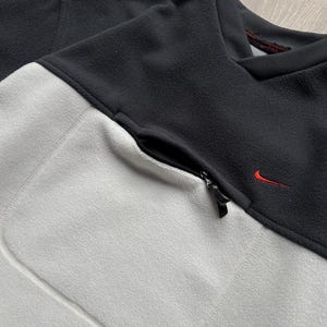 May include: A black and light grey fleece pullover with a zipped pocket and a small red Nike logo. The top has a crew neck and is made of a soft, textured material. The colour-blocked design adds a modern touch to this athletic-inspired garment.
