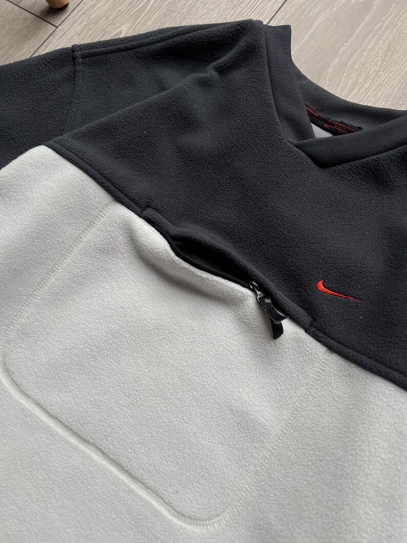 May include: A black and light grey fleece pullover with a zipped pocket and a small red Nike logo. The top has a crew neck and is made of a soft, textured material. The colour-blocked design adds a modern touch to this athletic-inspired garment.