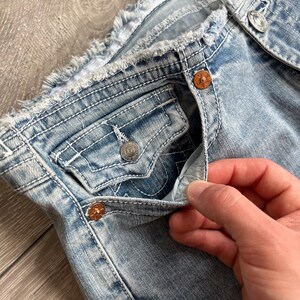 May include: Light blue denim jeans featuring a distressed, frayed waistband, copper-colored metal rivets, and a small coin pocket with a button flap. The fabric has a light wash finish with visible white contrast stitching throughout the garment.