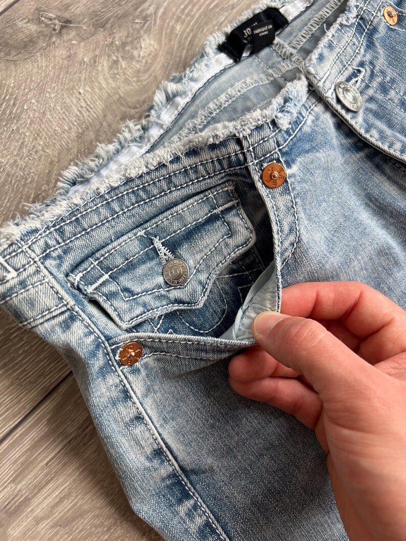 May include: Light blue denim jeans featuring a distressed, frayed waistband, copper-colored metal rivets, and a small coin pocket with a button flap. The fabric has a light wash finish with visible white contrast stitching throughout the garment.
