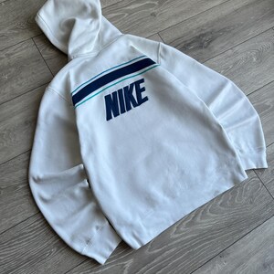 Puede incluir: Sudadera con capucha blanca con un dise&ntilde;o de rayas turquesas y azul marino en la espalda. La palabra "NIKE" est&aacute; bordada en azul marino. La sudadera tiene capucha y mangas largas.