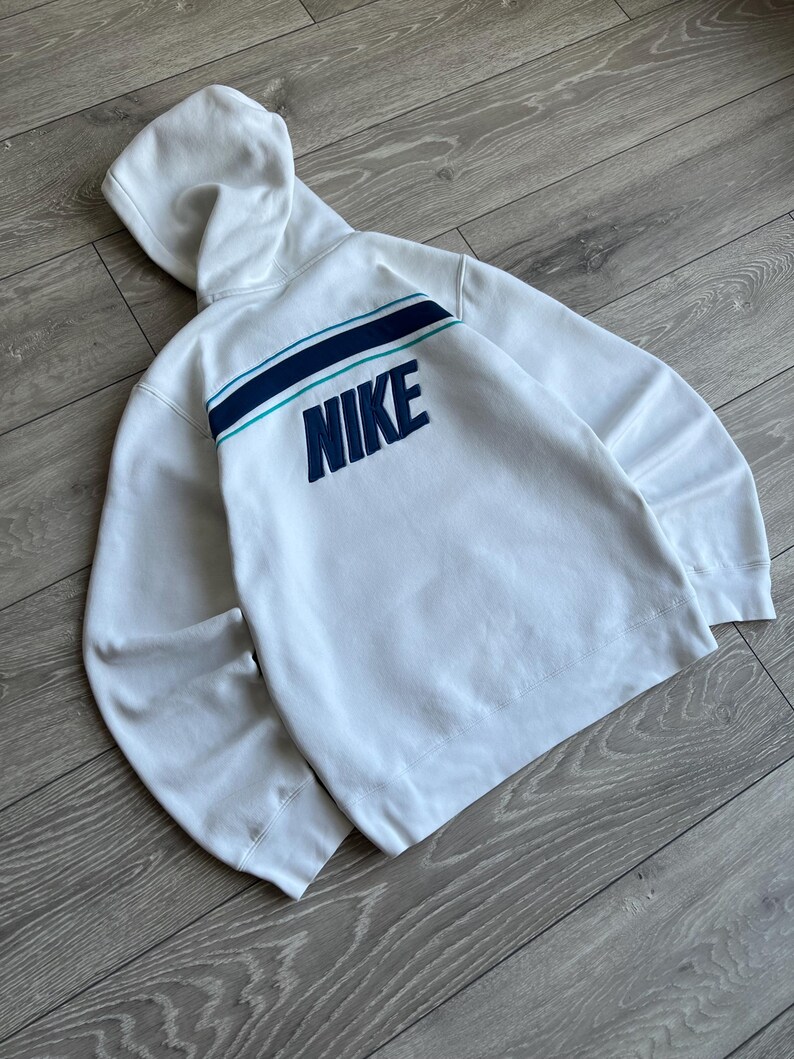 Puede incluir: Sudadera con capucha blanca con un dise&ntilde;o de rayas turquesas y azul marino en la espalda. La palabra "NIKE" est&aacute; bordada en azul marino. La sudadera tiene capucha y mangas largas.