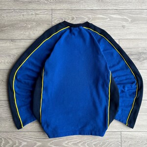 May include: A blue sweatshirt with black sleeves and yellow trim. The crew neck sweatshirt has a solid blue front and back. The sleeves are black with a yellow stripe that runs along the top of the sleeves.