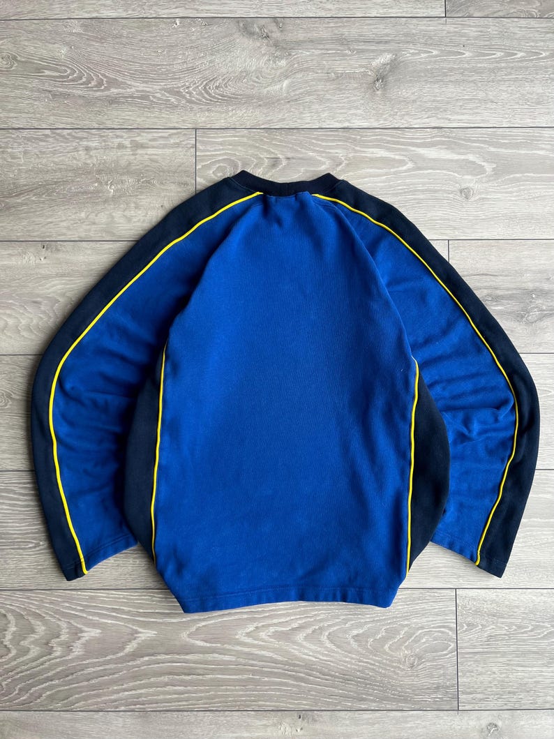 May include: A blue sweatshirt with black sleeves and yellow trim. The crew neck sweatshirt has a solid blue front and back. The sleeves are black with a yellow stripe that runs along the top of the sleeves.