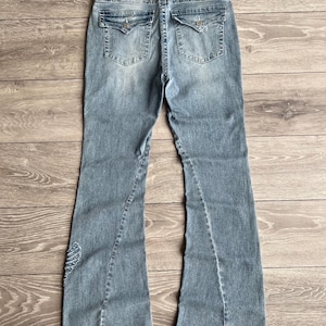 May include: Light blue denim jeans featuring a flared leg silhouette, a frayed waistband, and two back pockets with button-flap closures. The denim has a faded wash, visible stitching details, and a small embroidered wing design on the lower leg.