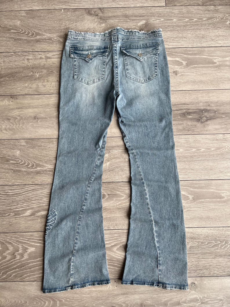 May include: Light blue denim jeans featuring a flared leg silhouette, a frayed waistband, and two back pockets with button-flap closures. The denim has a faded wash, visible stitching details, and a small embroidered wing design on the lower leg.