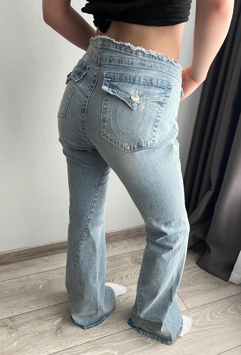May include: Light blue denim jeans featuring a frayed waistband, two back pockets with buttoned flaps, and prominent white stitching. The jeans have a bootcut silhouette with a faded wash, showcasing a classic five-pocket design and metal hardware.