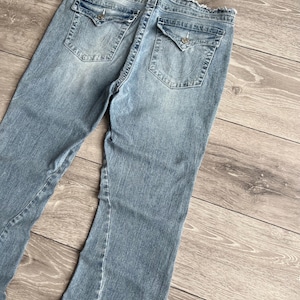 May include: Light blue denim jeans featuring a distressed, frayed waistband edge. The back includes two flap pockets with silver-toned button closures and a stitched horseshoe-style logo detail on each pocket. The denim has a faded wash finish.