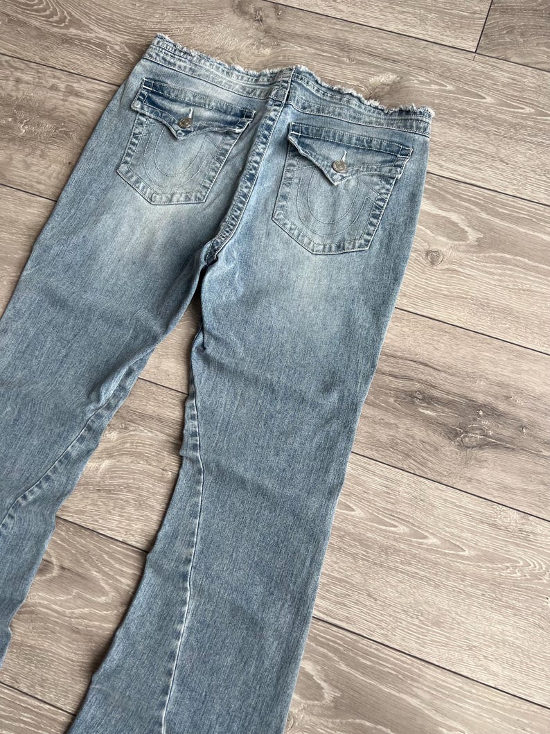 May include: Light blue denim jeans featuring a distressed, frayed waistband edge. The back includes two flap pockets with silver-toned button closures and a stitched horseshoe-style logo detail on each pocket. The denim has a faded wash finish.
