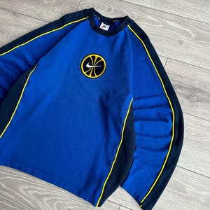 May include: Blue Nike sweatshirt with a basketball logo. The sweatshirt has black and yellow accents along the sleeves and neckline. The Nike logo is visible on the chest. The sweatshirt is made of a soft material.