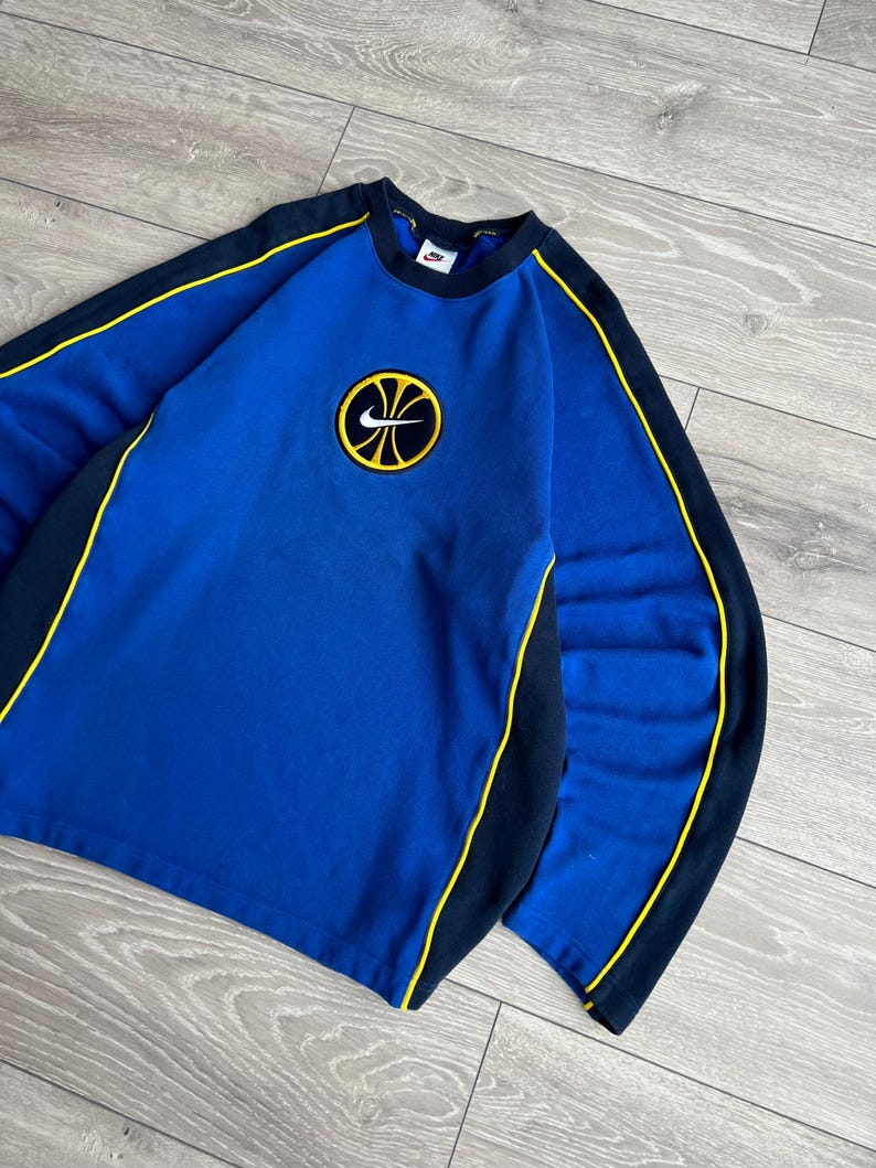 May include: Blue Nike sweatshirt with a basketball logo. The sweatshirt has black and yellow accents along the sleeves and neckline. The Nike logo is visible on the chest. The sweatshirt is made of a soft material.