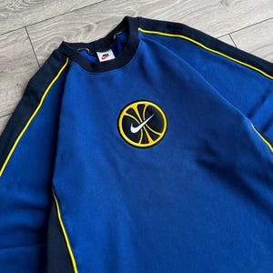 May include: Blue sweatshirt with black and yellow accents. Features a circular patch with a yellow basketball design and a white Nike swoosh logo. The sweatshirt has a crew neck and long sleeves.