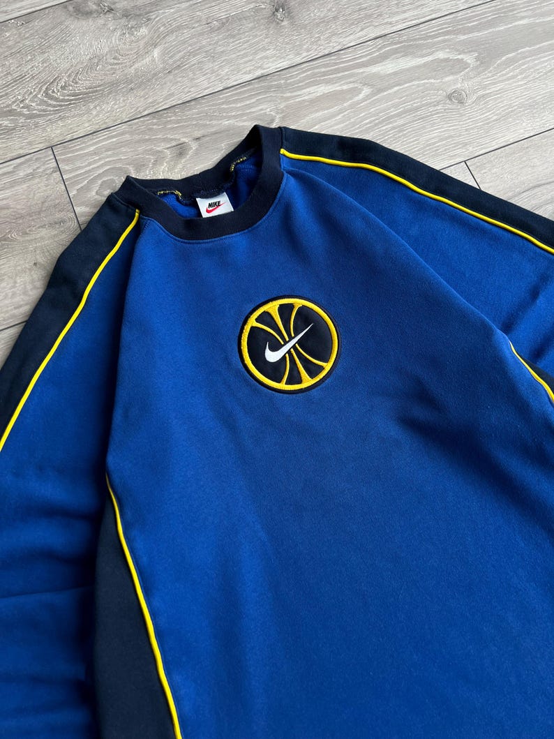May include: Blue sweatshirt with black and yellow accents. Features a circular patch with a yellow basketball design and a white Nike swoosh logo. The sweatshirt has a crew neck and long sleeves.