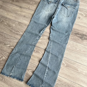 May include: Light blue denim jeans featuring a flared leg silhouette, a frayed waistband, and two back pockets with button-down flaps. The denim has a faded wash effect, visible stitching details, and a small embroidered wing design on the lower leg.
