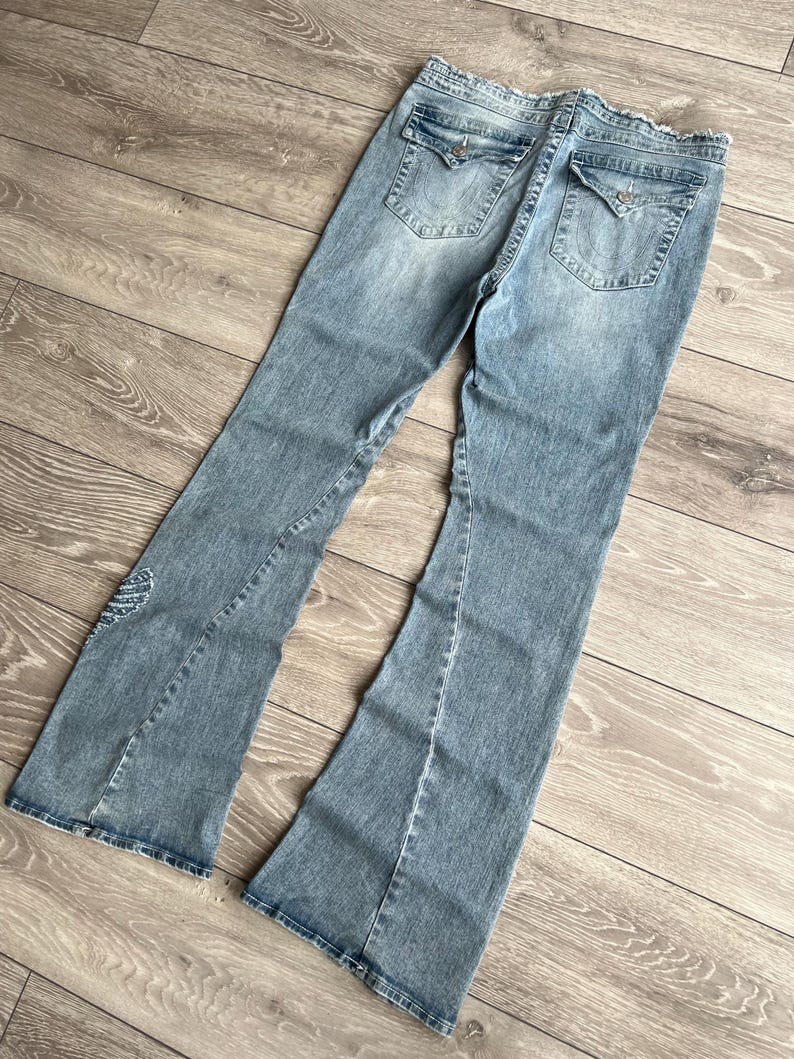 May include: Light blue denim jeans featuring a flared leg silhouette, a frayed waistband, and two back pockets with button-down flaps. The denim has a faded wash effect, visible stitching details, and a small embroidered wing design on the lower leg.