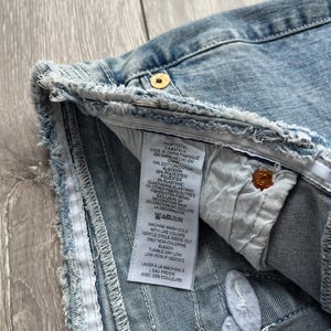 May include: A light blue denim garment with a frayed, raw-edge hem and a white interior care label. The label lists a material composition of 70% cotton, 26% polyester, 2% elastane, and 2% other fibers, with instructions for machine washing.
