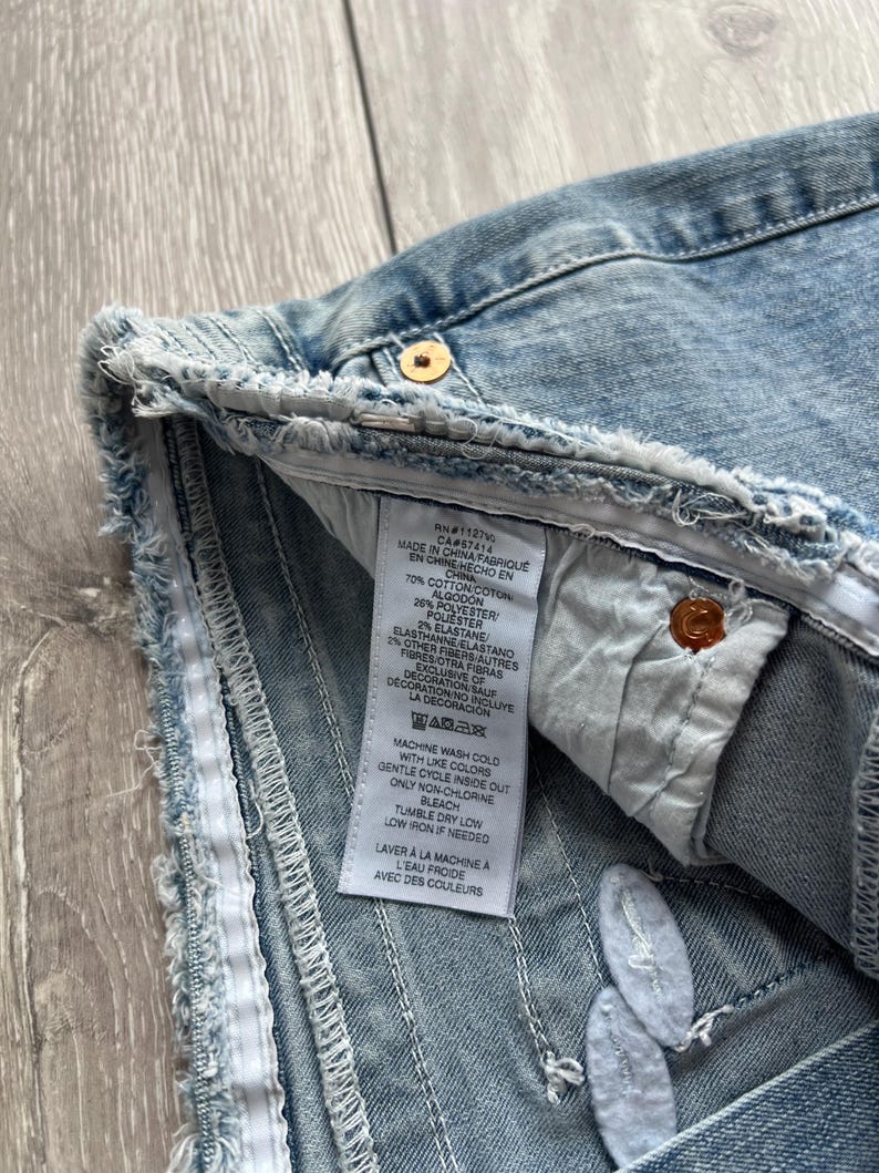 May include: A light blue denim garment with a frayed, raw-edge hem and a white interior care label. The label lists a material composition of 70% cotton, 26% polyester, 2% elastane, and 2% other fibers, with instructions for machine washing.