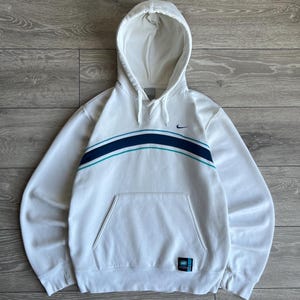 Puede incluir: Sudadera con capucha blanca con bolsillo delantero y dise&ntilde;o de rayas azules y turquesas en el pecho. Cuenta con un peque&ntilde;o logotipo de Nike y una etiqueta en la parte inferior derecha. La sudadera est&aacute; hecha de un material suave.