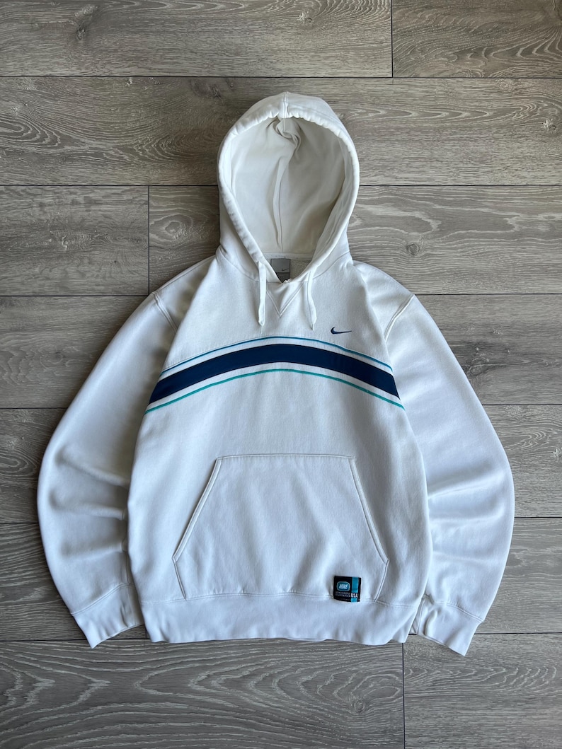 Puede incluir: Sudadera con capucha blanca con bolsillo delantero y dise&ntilde;o de rayas azules y turquesas en el pecho. Cuenta con un peque&ntilde;o logotipo de Nike y una etiqueta en la parte inferior derecha. La sudadera est&aacute; hecha de un material suave.