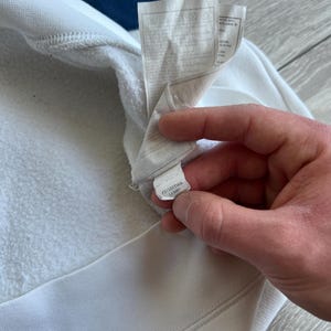 Puede incluir: Primer plano de una sudadera blanca con un dise&ntilde;o de rayas azules y marino en el cuello. La etiqueta es visible, con instrucciones de cuidado y un c&oacute;digo de producto. La tela parece suave y texturizada.