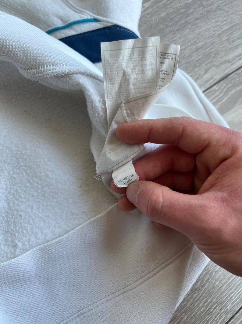 Puede incluir: Primer plano de una sudadera blanca con un dise&ntilde;o de rayas azules y marino en el cuello. La etiqueta es visible, con instrucciones de cuidado y un c&oacute;digo de producto. La tela parece suave y texturizada.