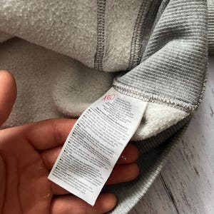 Puede incluir: Primer plano del interior de una sudadera gris con un forro blanco y difuso. Una etiqueta con texto en varios idiomas es visible. El exterior de la sudadera es de tejido de punto acanalado. Se ve la mano que sostiene la sudadera.